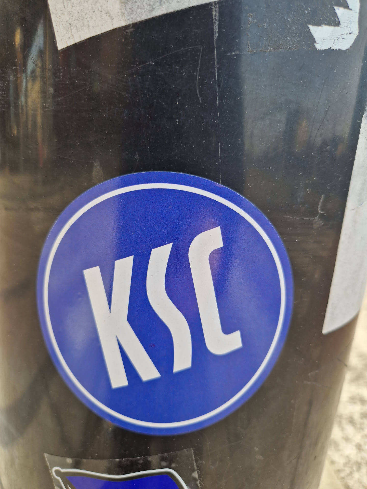 A round blue sticker with the letters KSC in a stylized sans-serif font. The sticker is affixed to a dark-colored pole.