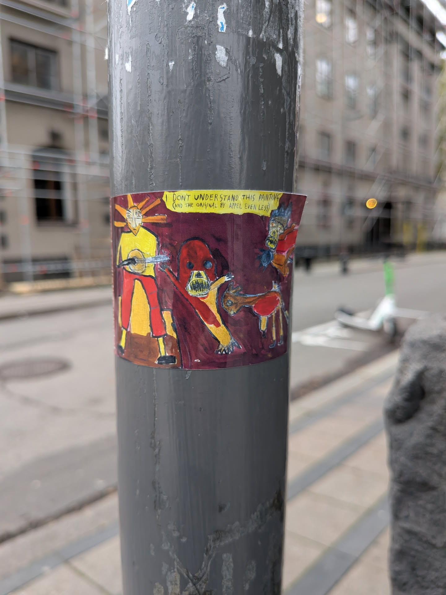 The sticker features a colorful, hand-drawn illustration with a dark maroon background. In the center, a stylized figure with a yellow body, red pants, and a mask-like face with orange rays emanating from its head is depicted playing a banjo. To the right, a red, monstrous creature with sharp teeth and a distorted face lunges forward, with a smaller, brown, deer-like creature in front of it. The overall style is crude and cartoonish, with bold outlines and vibrant colors. A yellow banner runs across the top of the sticker with text.