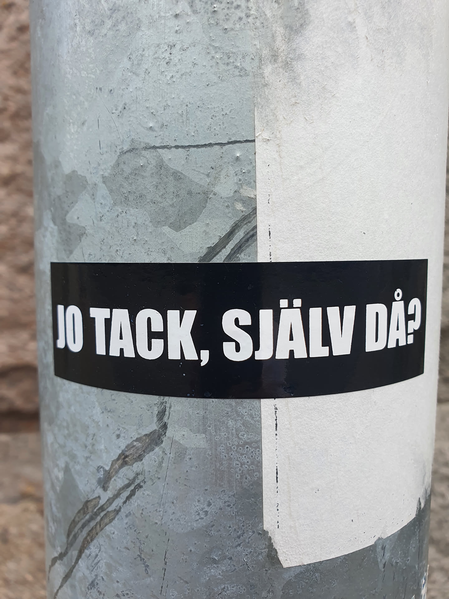 A black sticker on a gray pole with Swedish text.  The text translates roughly to "Yeah thanks, yourself then?" It's a common informal response in Swedish conversations.