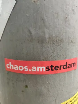 A red and white sticker with the text 'chaos.amsterdam' on it. It's stuck on a grey pole.