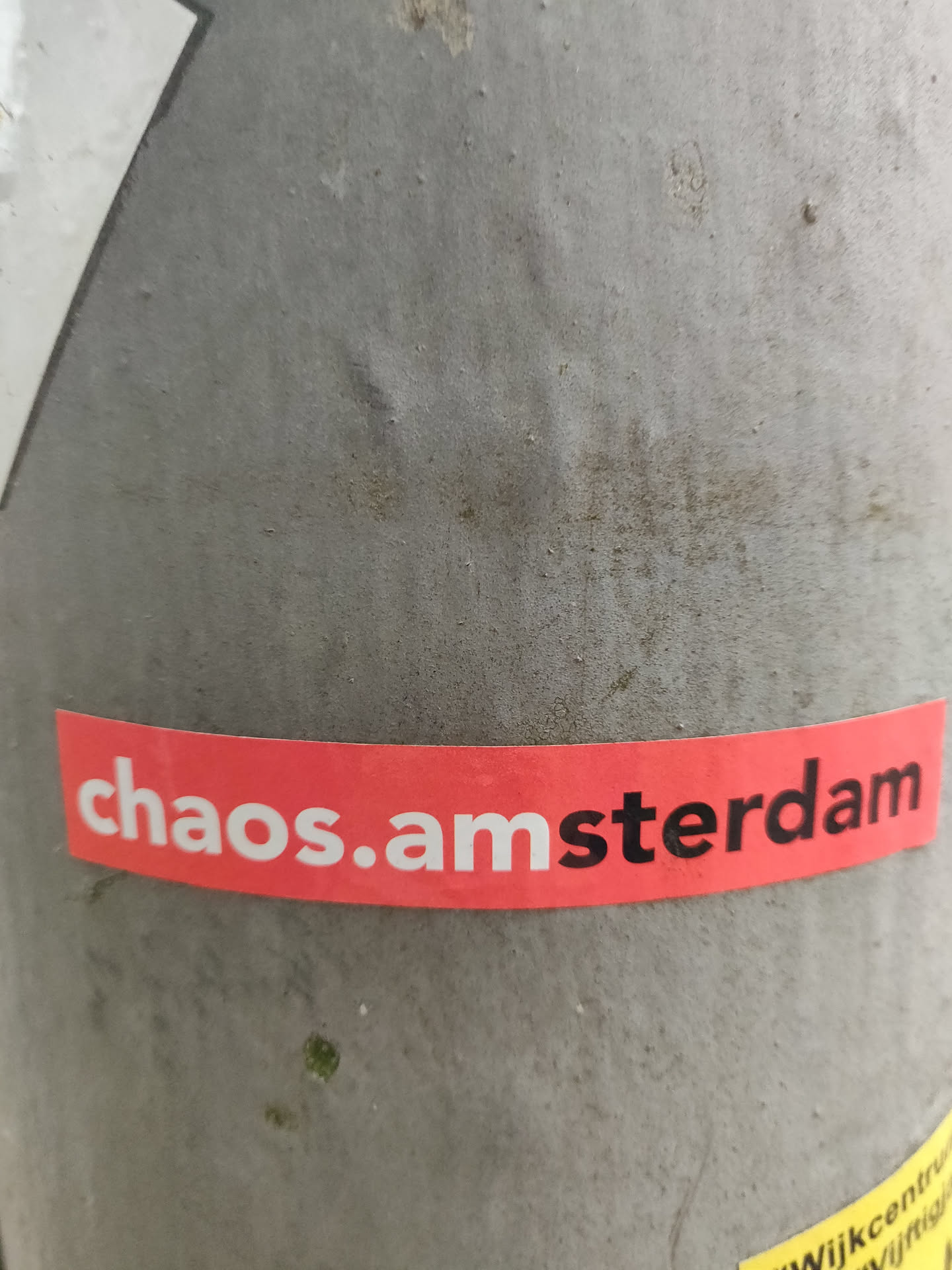 A red and white sticker with the text 'chaos.amsterdam' on it. It's stuck on a grey pole.