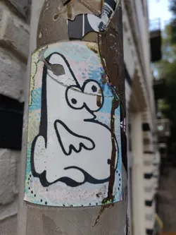 A charming sticker featuring a simple, cartoon-like creature in black and white with a pastel background. The sticker shows some wear, suggesting it has been exposed to the elements. It is affixed to a weathered pillar or pole, adding to its urban aesthetic.