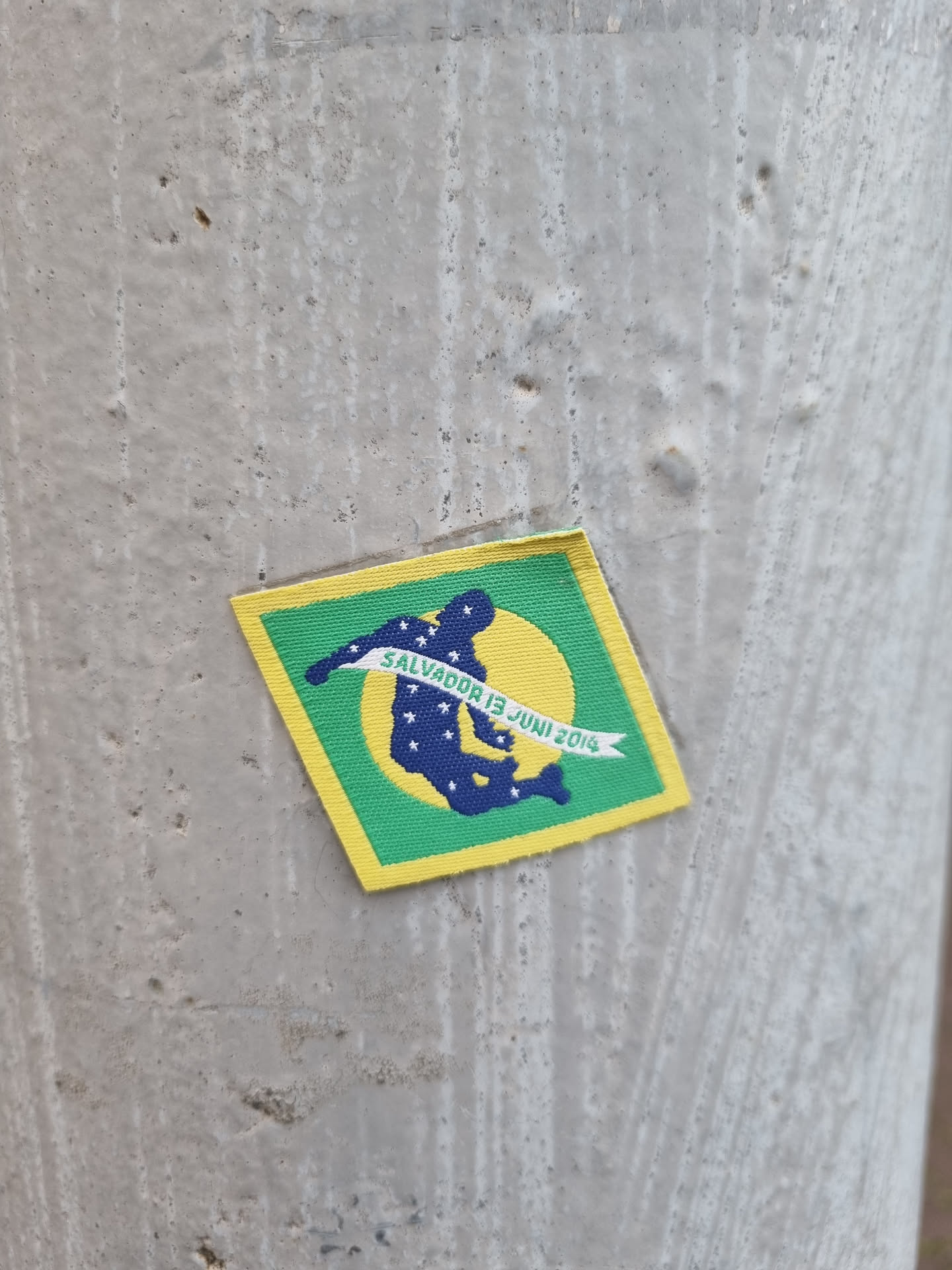 A small, diamond-shaped sticker featuring a stylized figure against a yellow sun and green background. The text 'Salvador 13 Juni 2014' is visible. The style suggests a commemorative or event-related sticker.