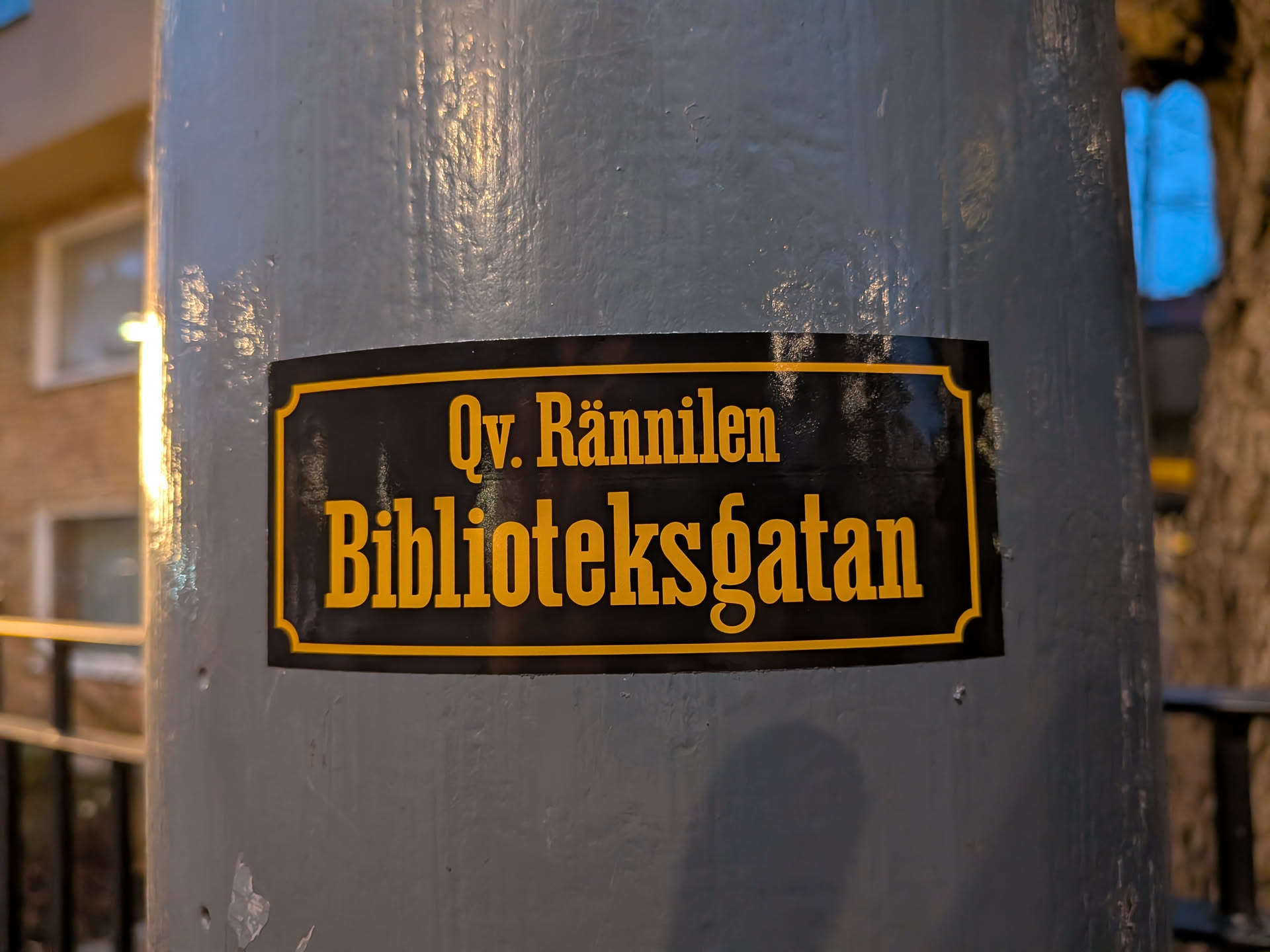 The sticker is rectangular with rounded corners and a decorative border. It features a black background with bold, yellow text. The top line reads 'Qv. Rännilen' and the second line reads 'Biblioteksgatan'. The text is in a serif font, and the overall design resembles a traditional street sign.