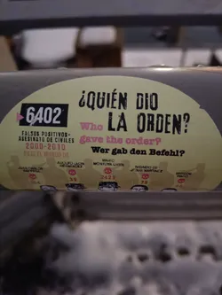 This sticker features the question "¿Quién dio la orden?" ("Who gave the order?") in Spanish, with English and German translations.  It also lists names and numbers, possibly referring to individuals involved in the events. The graphic design is stark and impactful. 