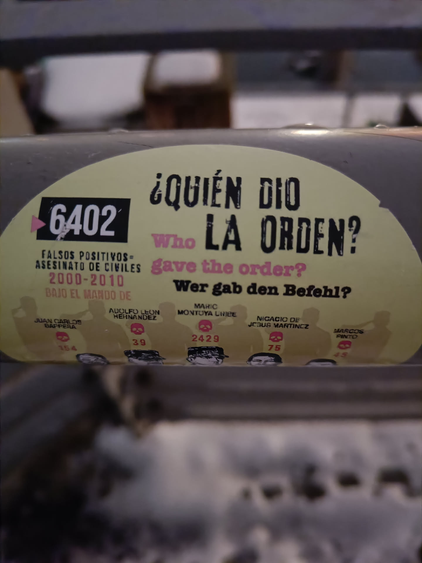 This sticker features the question "¿Quién dio la orden?" ("Who gave the order?") in Spanish, with English and German translations.  It also lists names and numbers, possibly referring to individuals involved in the events. The graphic design is stark and impactful. 