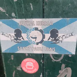 A sticker featuring a flag-like design with the text "EUSKAL HERRIA" at the top and "SANKT PAULI FANS" at the bottom. The central design includes a gear and a laurel wreath, along with stylized figures holding weapons. A smaller sticker is visible below the main sticker that says "Freund der Helfer".