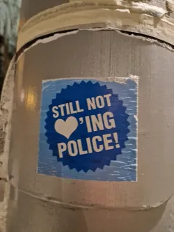 A blue sticker with a heart and the text "Still not loving police!" is attached to a pole. The sticker has a slightly worn appearance.