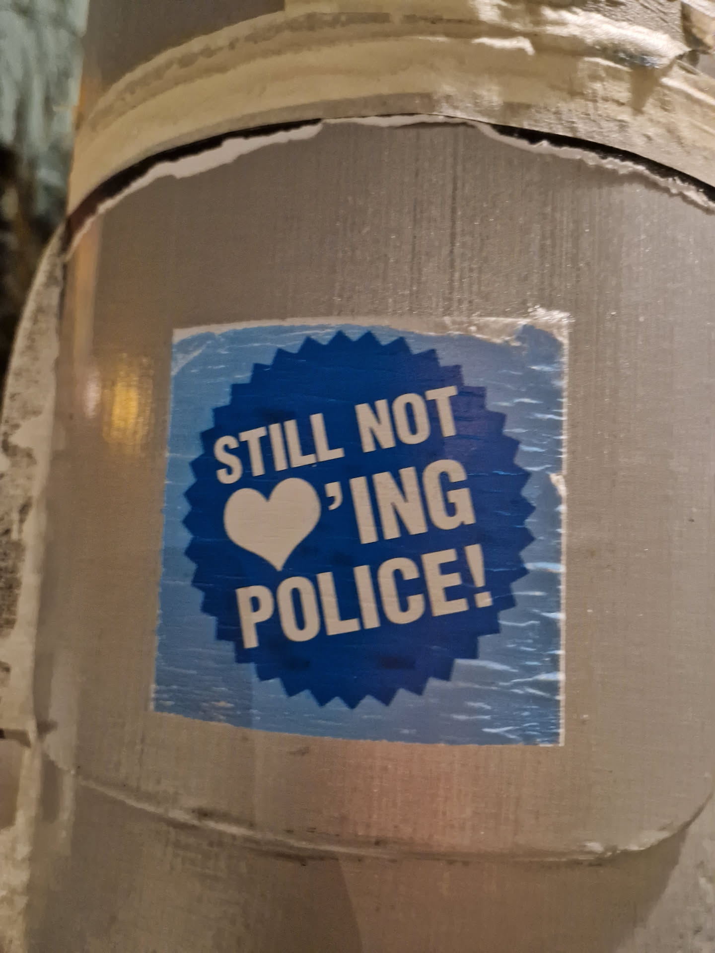 A blue sticker with a heart and the text "Still not loving police!" is attached to a pole. The sticker has a slightly worn appearance.