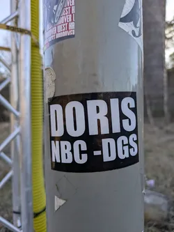 A black and white rectangular sticker with the text "DORIS NBC-DGS" is adhered to a gray metal pole. The sticker has a black background with the text in white block letters.