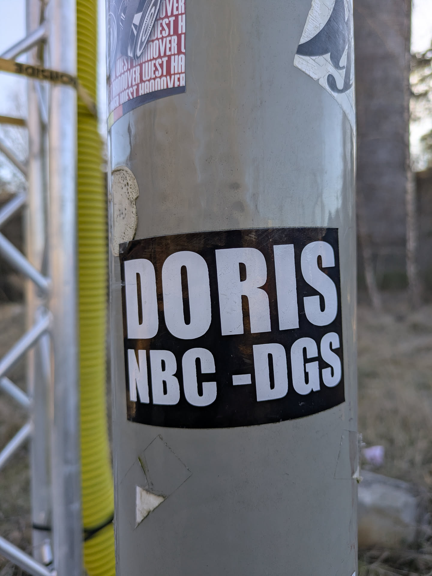 A black and white rectangular sticker with the text "DORIS NBC-DGS" is adhered to a gray metal pole. The sticker has a black background with the text in white block letters.