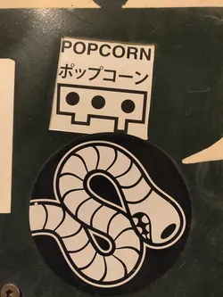 A black and white sticker of a worm design on a black circular background. Above it is a rectangular sticker with the word POPCORN written in English and Japanese.