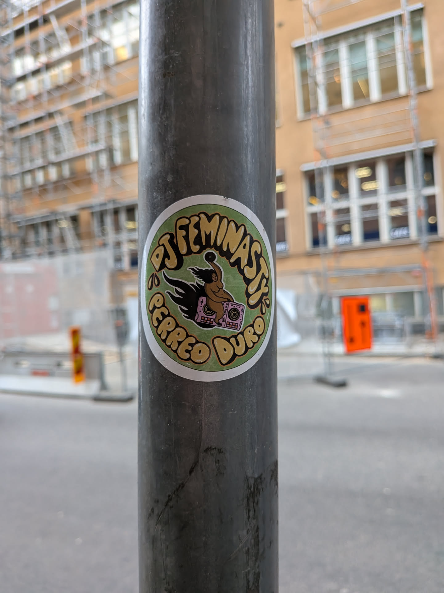 A circular sticker affixed to a grey metal street pole. It features a muted green background enclosed in a thin white circular border. At the center is an illustration of a woman with long black hair riding atop a pink boombox that emits black flames behind it, holding a vinyl record high above her head. Surrounding this central drawing are stylized, curvy beige letters with black drop shadows that read 'DJ FEMINASTY' around the top and 'PERREO DURO' along the bottom.