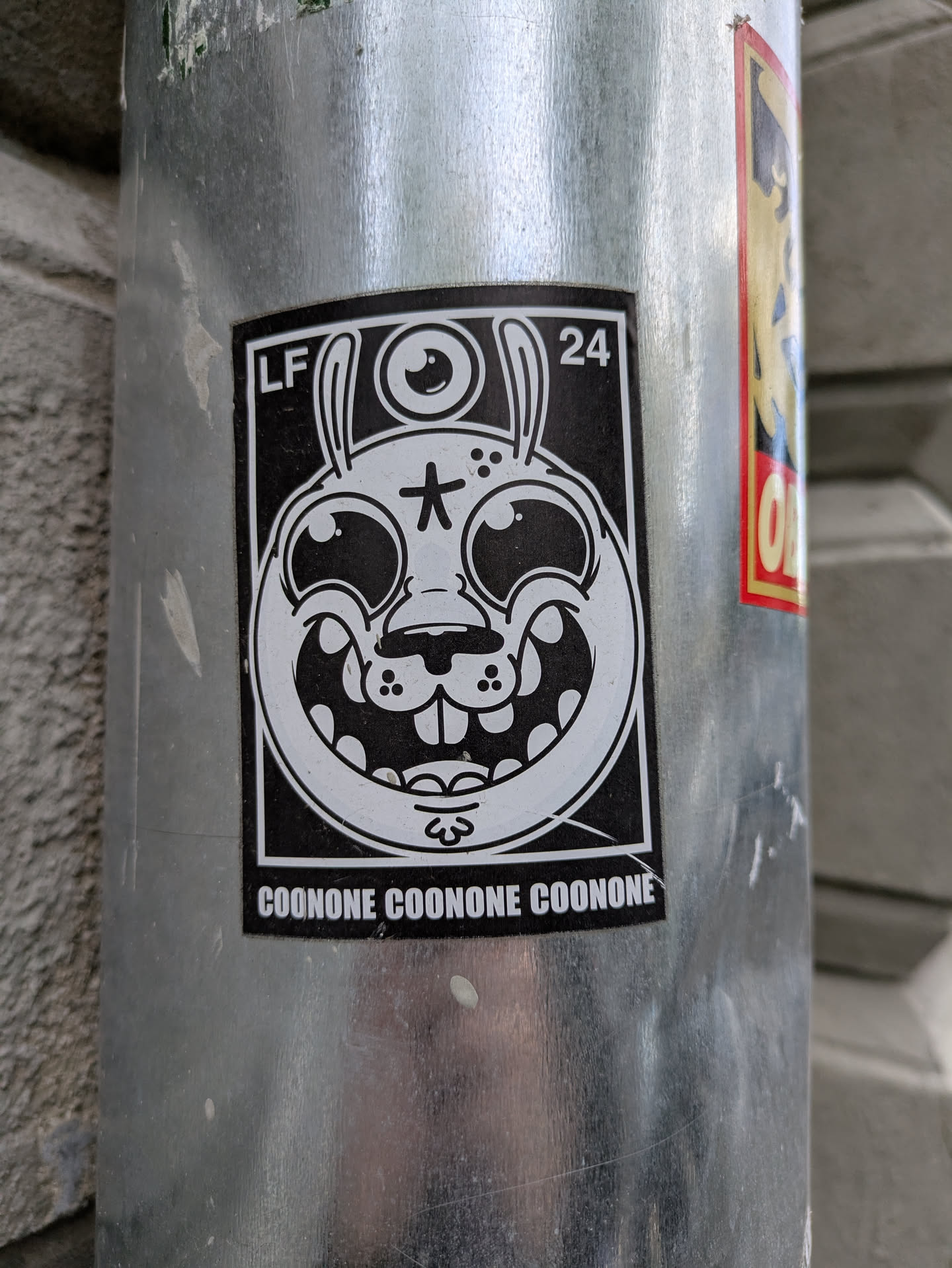 This sticker has a black background and white ink. The design is a cartoon-style rabbit head with exaggerated features. It has a third eye on its forehead, a star on its face, and a wide, toothy grin. The text "COONONE COONONE COONONE" is printed at the bottom of the sticker. It is affixed to a silver pole.