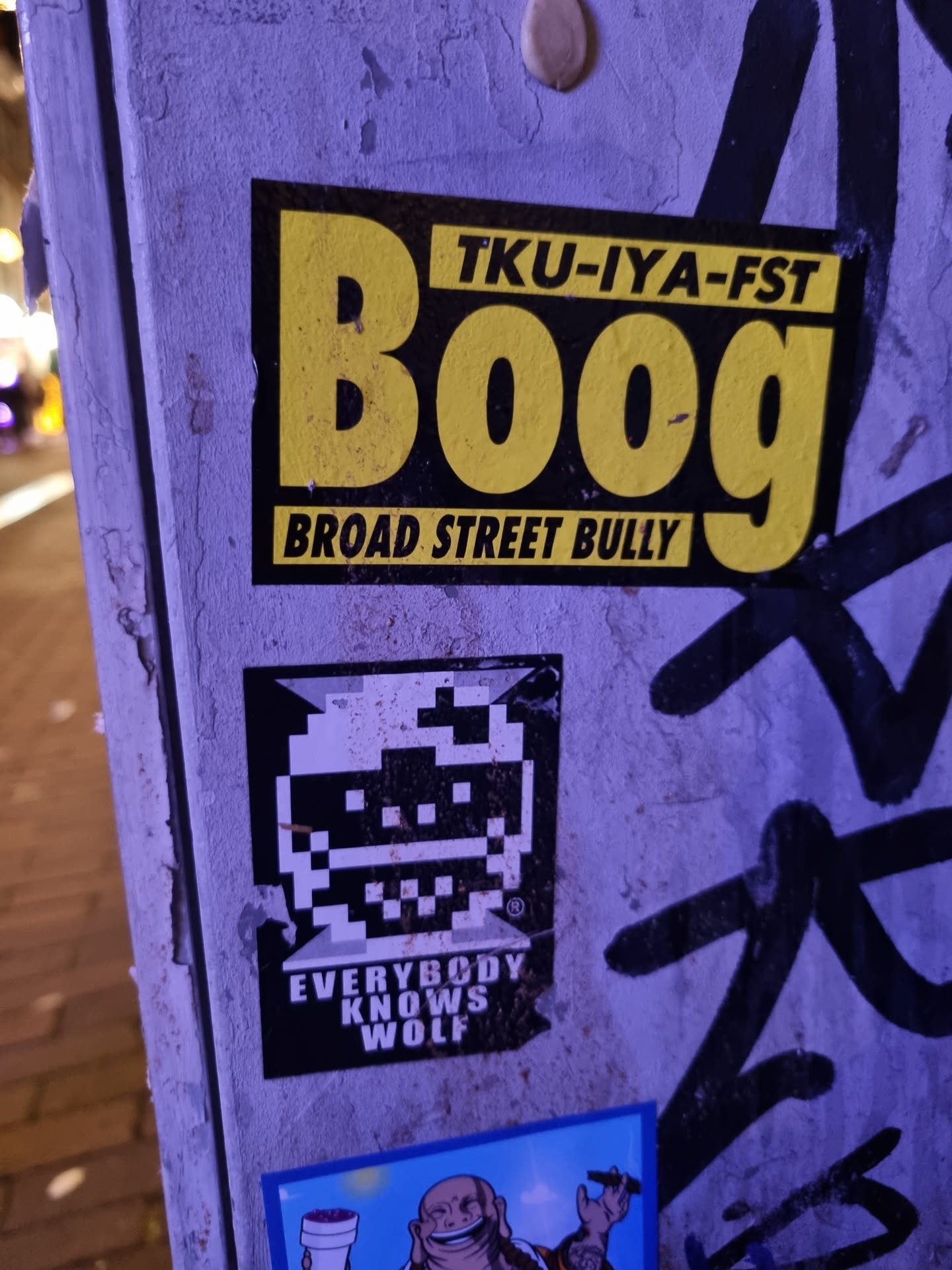 A yellow and black sticker with the text "Tku-iya-fst Boog" and below it "Broad Street Bully". There is also a smaller pixel art sticker underneath it with the text "Everybody Knows Wolf".