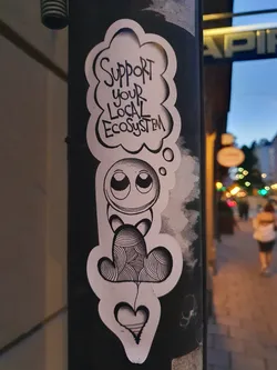 Black and white illustration sticker featuring a cute cartoon character with a thought bubble that says "Support your local ecosystem". The character is holding a heart-shaped object. 