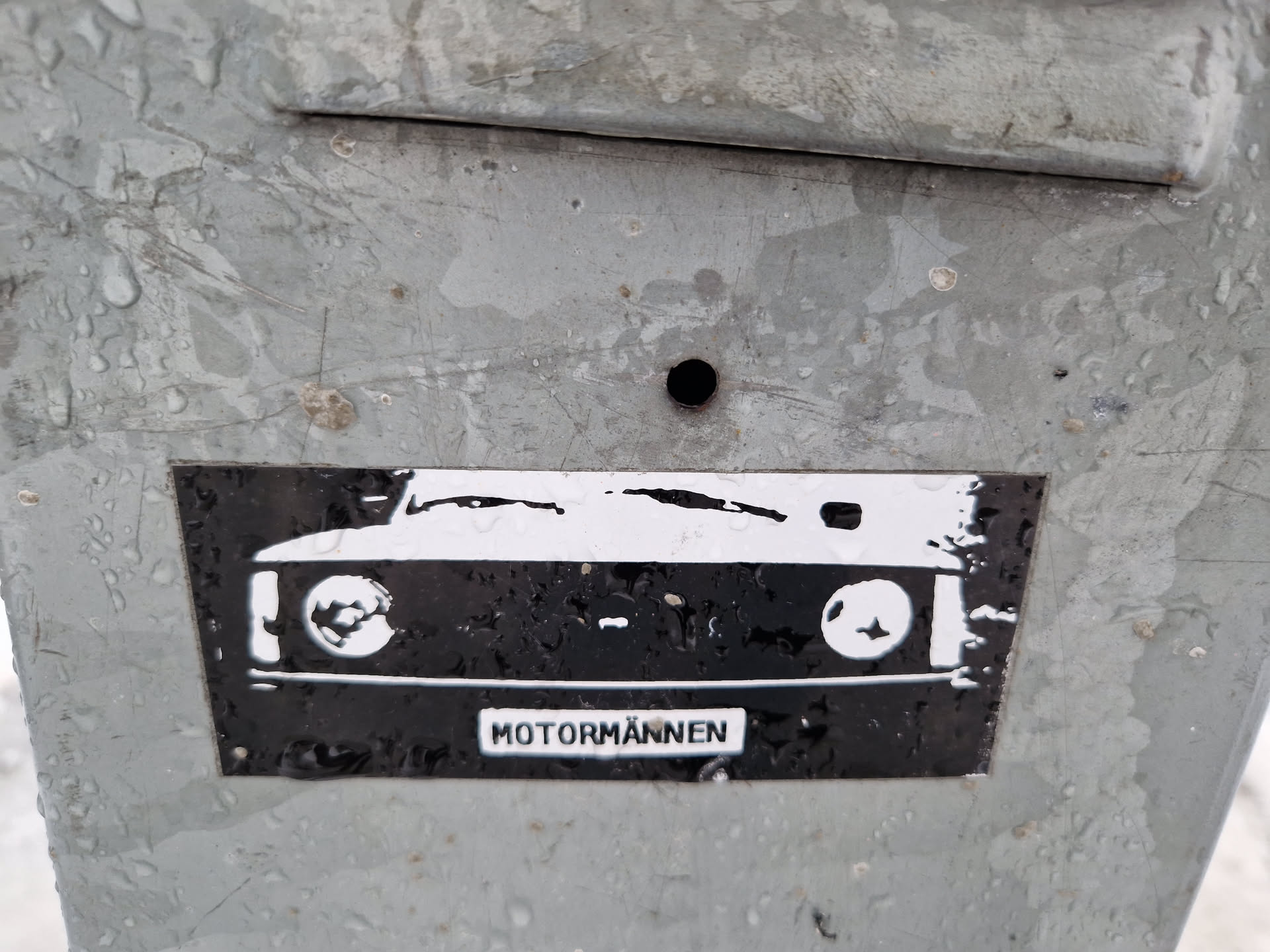 A black and white sticker featuring the front of a classic car. The word "MOTORMANNEN" is visible at the bottom. The sticker is affixed to a weathered grey metal surface.
