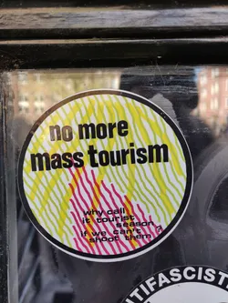 Round sticker with a design of yellow and pink lines.  The text on the sticker says: "no more mass tourism" and "why call it tourist season if we can't shoot them?"