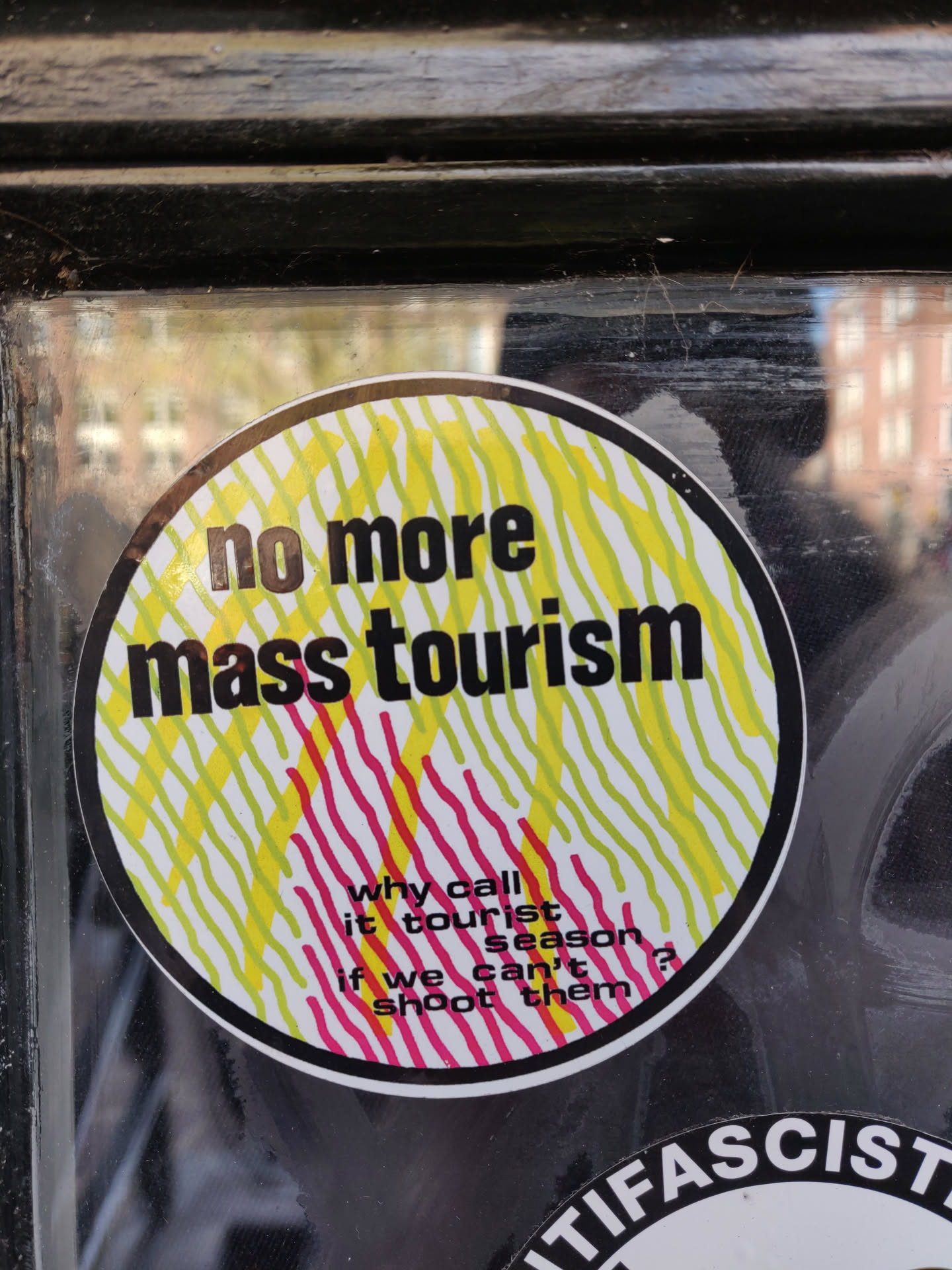 Round sticker with a design of yellow and pink lines.  The text on the sticker says: "no more mass tourism" and "why call it tourist season if we can't shoot them?"