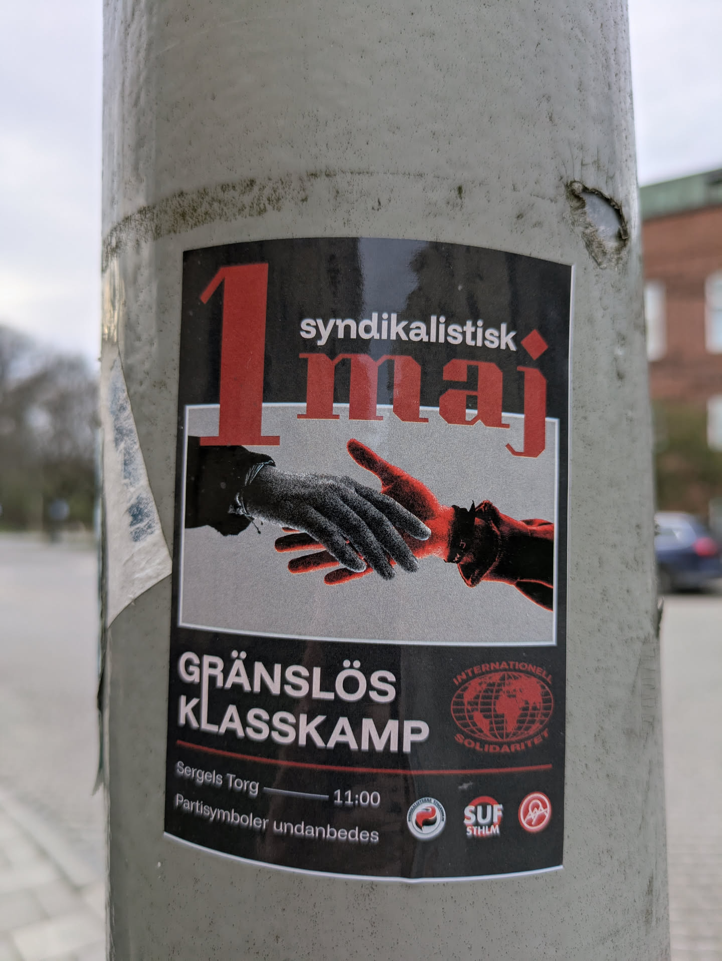 A sticker on a pole featuring an image of two hands reaching for each other, one in black and white and the other in red. The text reads "1 syndikalistisk maj" at the top, and "GRÄNSLÖS KLASSKAMP" below the hands. The sticker also includes the location and time: "Sergels Torg 11:00", with a note "Partisymboler undanbedes" and the symbol of "INTERNATIONAL SOLIDARITY", along with the SUF logo.