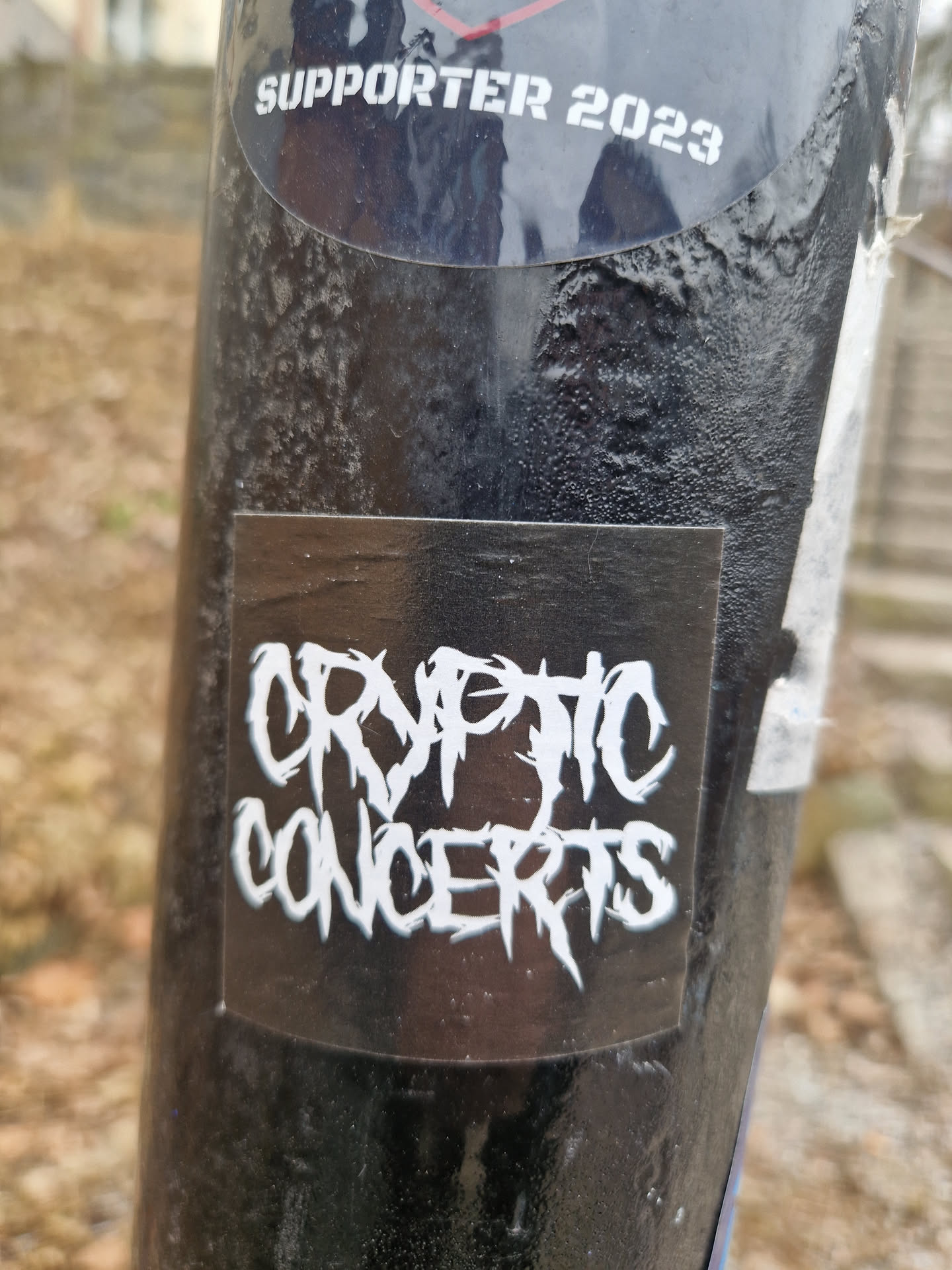 A black sticker with white text reading "Cryptic Concerts" in a stylized font. The sticker is affixed to a dark-colored pole.