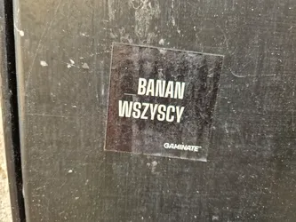 A black sticker with the text BANAN WSZYSCY and the word GAMIMATE at the bottom. The sticker is attached to a dark gray surface.