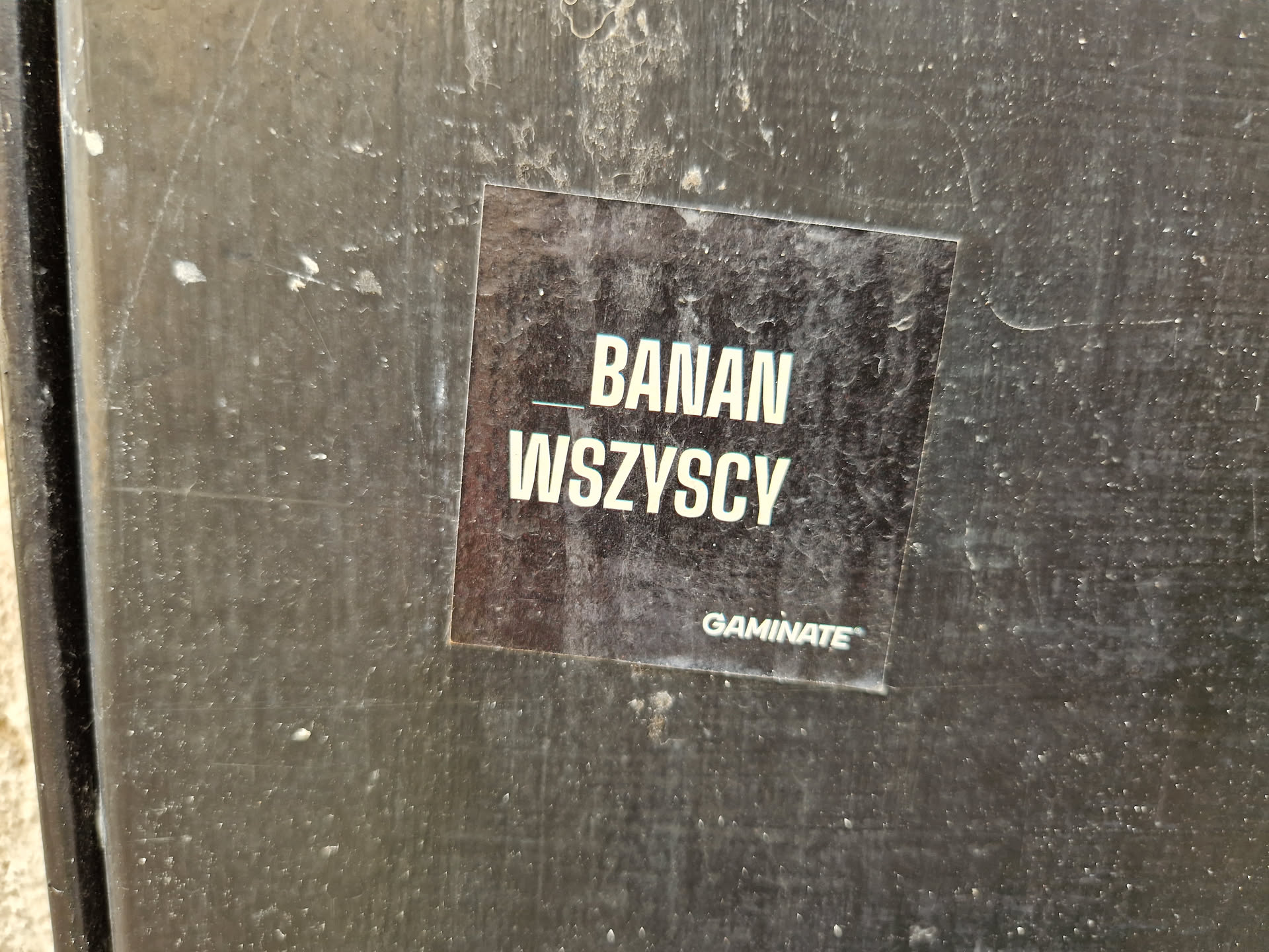 A black sticker with the text BANAN WSZYSCY and the word GAMIMATE at the bottom. The sticker is attached to a dark gray surface.