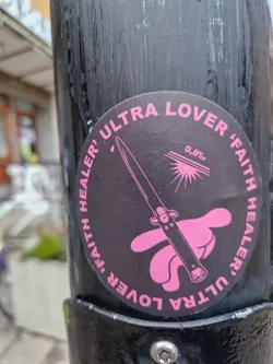 Round black and pink sticker with an illustration of a switchblade on a pink hand. The text "Ultra lover, faith healer" is written around the circle, with "0,8%" in the center.