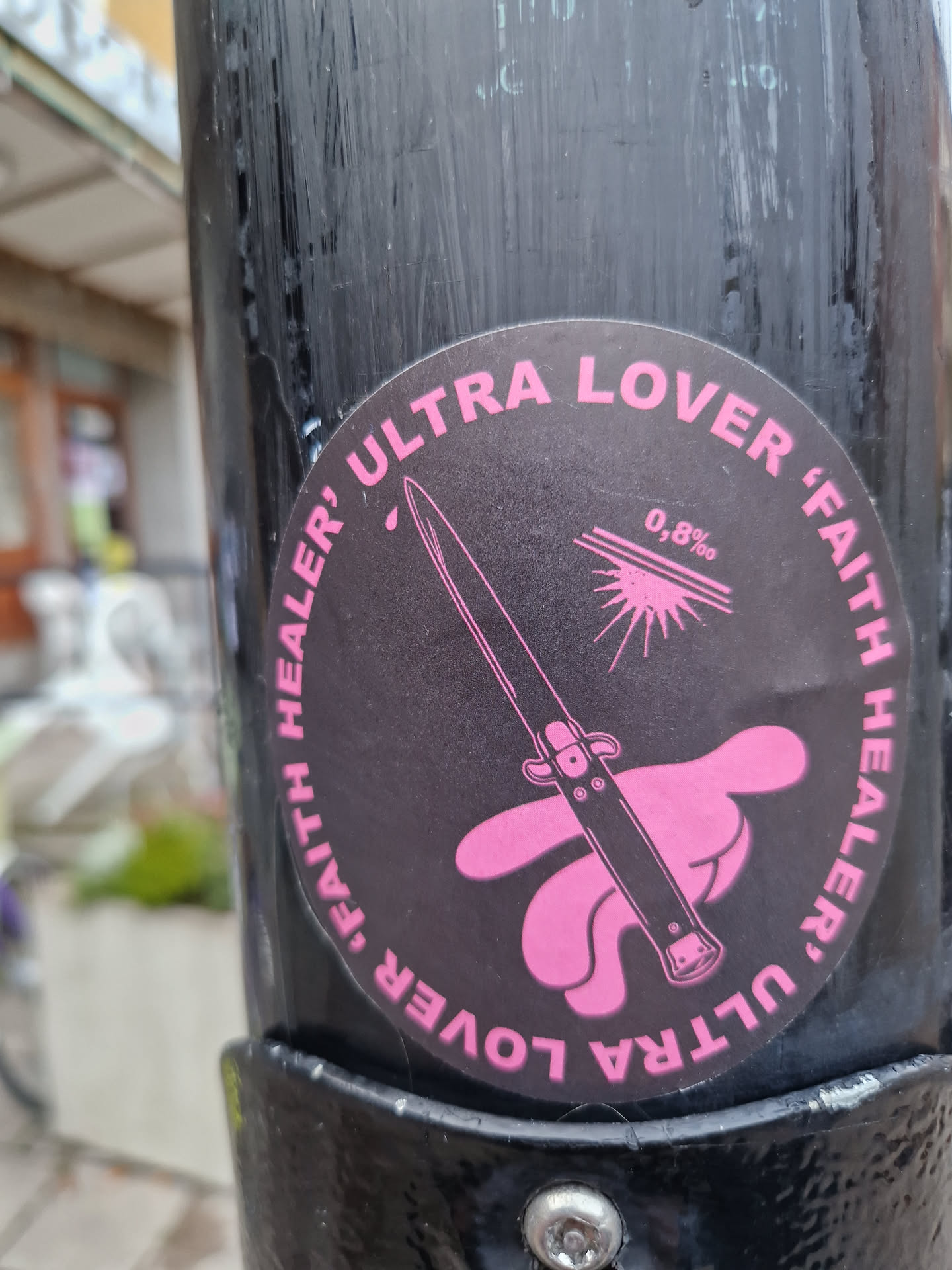 Round black and pink sticker with an illustration of a switchblade on a pink hand. The text "Ultra lover, faith healer" is written around the circle, with "0,8%" in the center.
