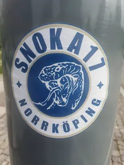 Close-up of a round sticker featuring the text "SNÖKA17 NORRKÖPING" arranged circularly around a central image of what seems to be a stylized brain or similar organic shape. The sticker has a white outer ring and a blue inner circle where the image and text reside.