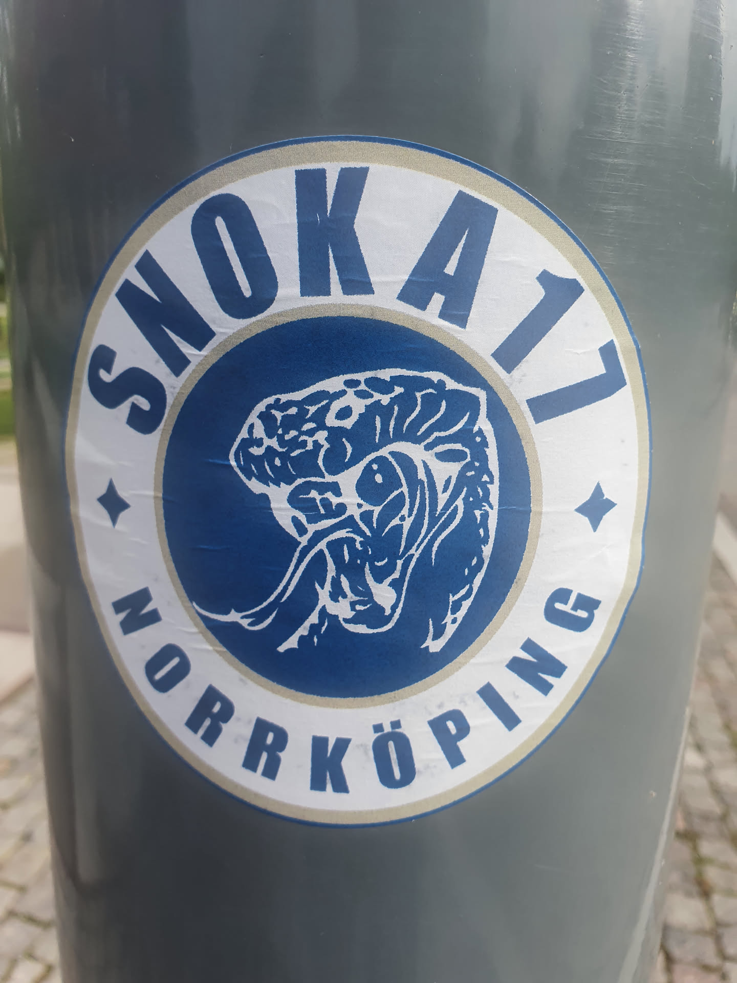 Close-up of a round sticker featuring the text "SNÖKA17 NORRKÖPING" arranged circularly around a central image of what seems to be a stylized brain or similar organic shape. The sticker has a white outer ring and a blue inner circle where the image and text reside.