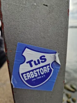 A blue and white sticker featuring the logo for TuS Erbstorf von 1965 e.V., a German sports club. The logo is a shield with the club's name and founding year. The sticker is peeling slightly at one corner and is affixed to a gray surface, possibly a metal pole or post, near a body of water.