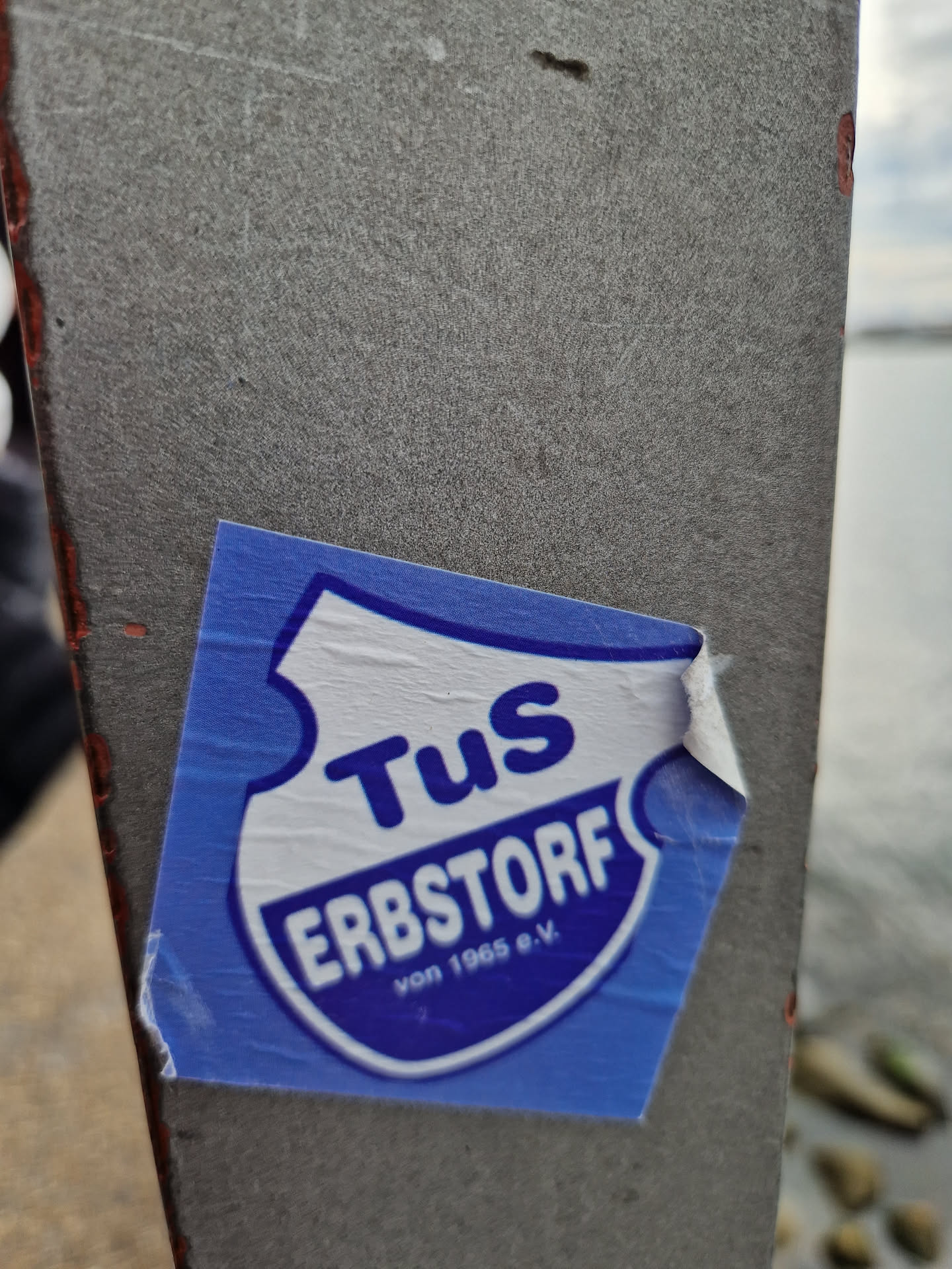 A blue and white sticker featuring the logo for TuS Erbstorf von 1965 e.V., a German sports club. The logo is a shield with the club's name and founding year. The sticker is peeling slightly at one corner and is affixed to a gray surface, possibly a metal pole or post, near a body of water.
