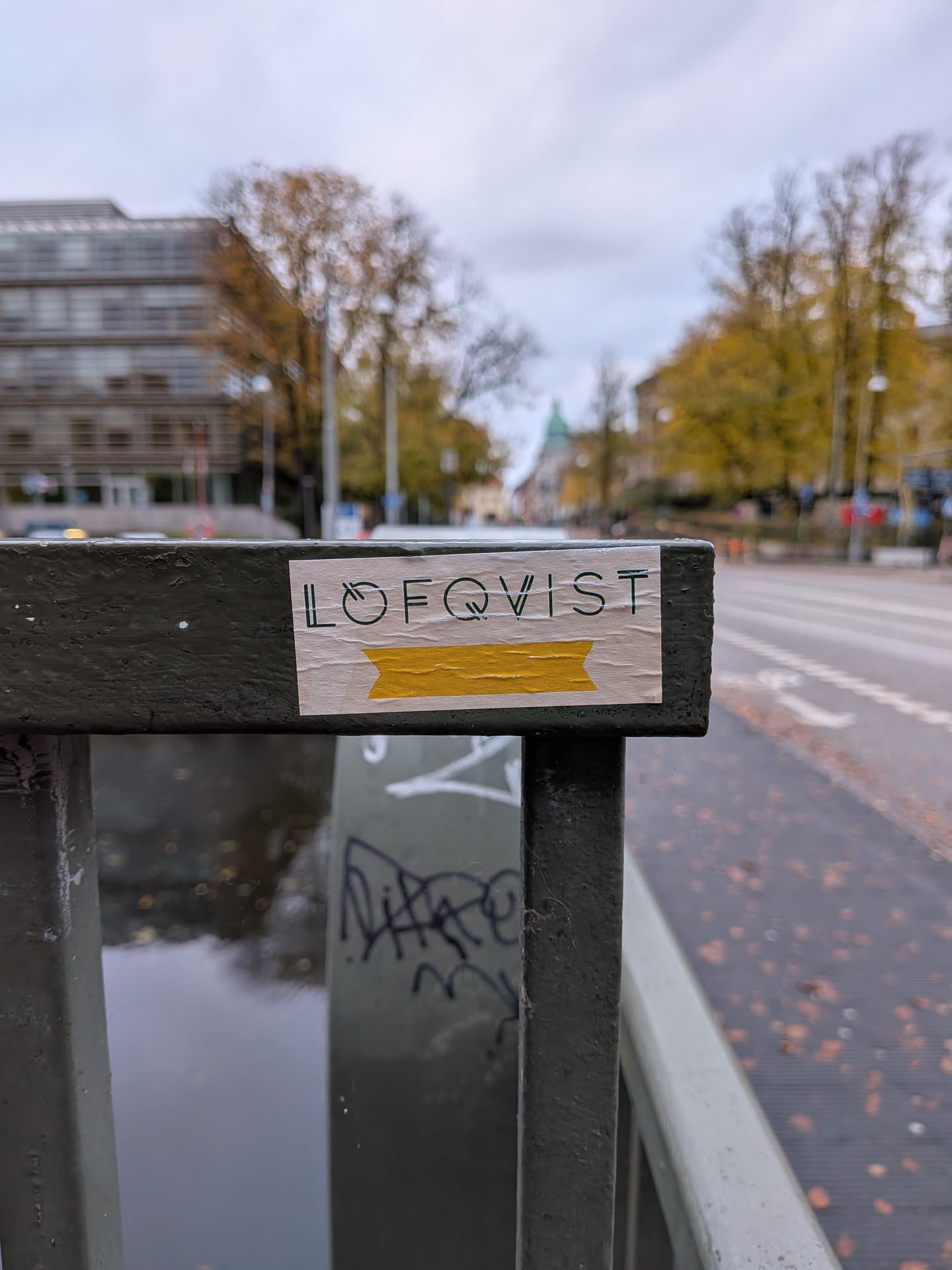 The sticker is rectangular with a light-colored background, appearing to be made of paper. It is affixed to a dark green metal railing. The top portion of the sticker features the word 'LOFQVIST' in a clean, sans-serif font with black lettering. The 'O' in 'LOFQVIST' is stylized to resemble a circle with a line through it, and the 'Q' has a similar circular element. Below the text, there is a horizontal band of bright yellow, resembling tape, with a pointed end on the right side. The edges of the sticker show signs of wear and slight peeling, indicating it has been placed outdoors.