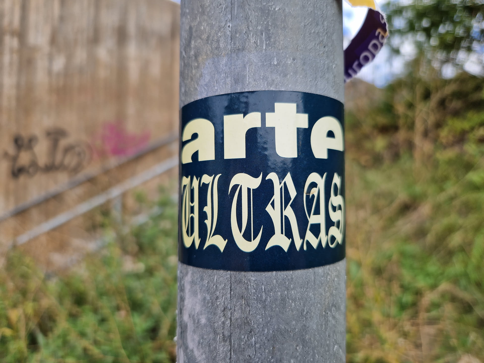 A dark blue sticker with the word \"arte\" in a sans-serif font and \"ULTRAS\" in a blackletter font. The sticker is attached to a grey metal pole outdoors. 