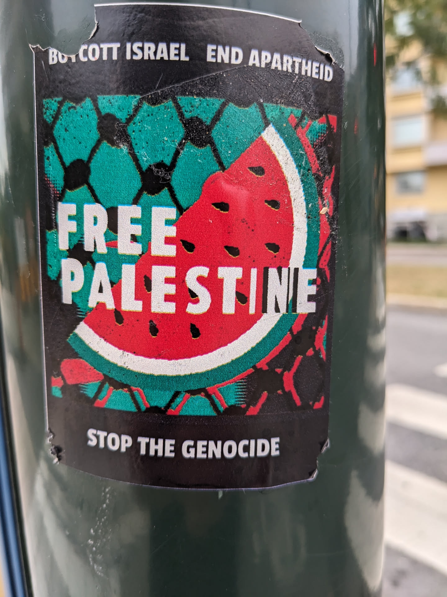 A sticker with a watermelon design split in half, one side representing Palestine with a teal background and the other side with red, representing Israel.  The text on the sticker says "Boycott Israel End Apartheid", "Free Palestine", and "Stop the Genocide".
