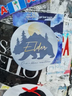 Round sticker featuring a bear silhouette, mountains, pine trees, and the word 'Elder' in gold lettering. The text at the bottom reads 'Once a plain mountain'.