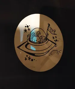 A round sticker features a hand-drawn illustration of a fluffy creature, possibly a rabbit, being abducted in a flying saucer.  The style is cartoonish and whimsical, with stars and other space-themed elements in the background. A small flag with a heart is attached to the UFO.