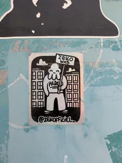 The sticker features a character standing between buildings, holding a flag that reads "ZEKO WAS". The character has "HERE" written on their chest.
