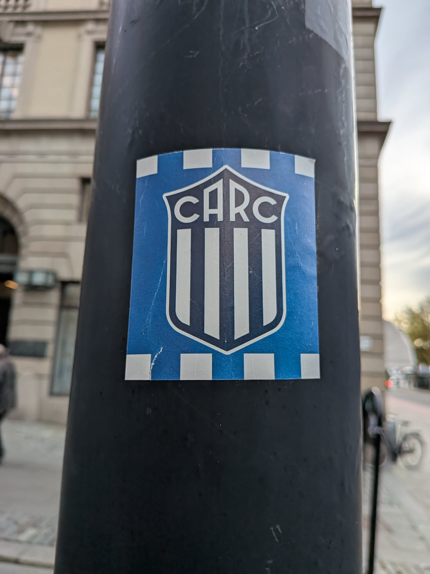 The sticker, affixed to a curved black metal pole, features a central shield emblem with the letters 'CARC' curved across the top over vertical stripes. The shield is set inside a blue rectangular field bordered on the top and bottom by alternating checkered blocks of light blue and white.
