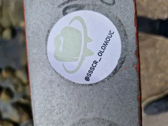 Round white sticker with a light green/grey illustration of what looks like a hat or helmet inside an orbit. The text '@SSSCR_OLOMOUC' is written below the illustration.
