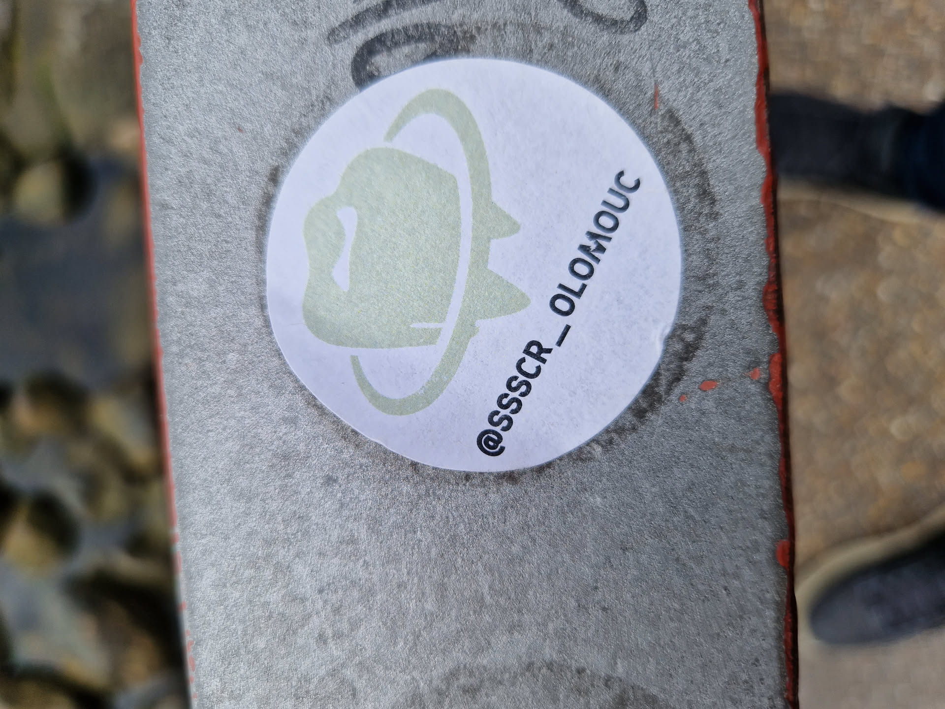Round white sticker with a light green/grey illustration of what looks like a hat or helmet inside an orbit. The text '@SSSCR_OLOMOUC' is written below the illustration.