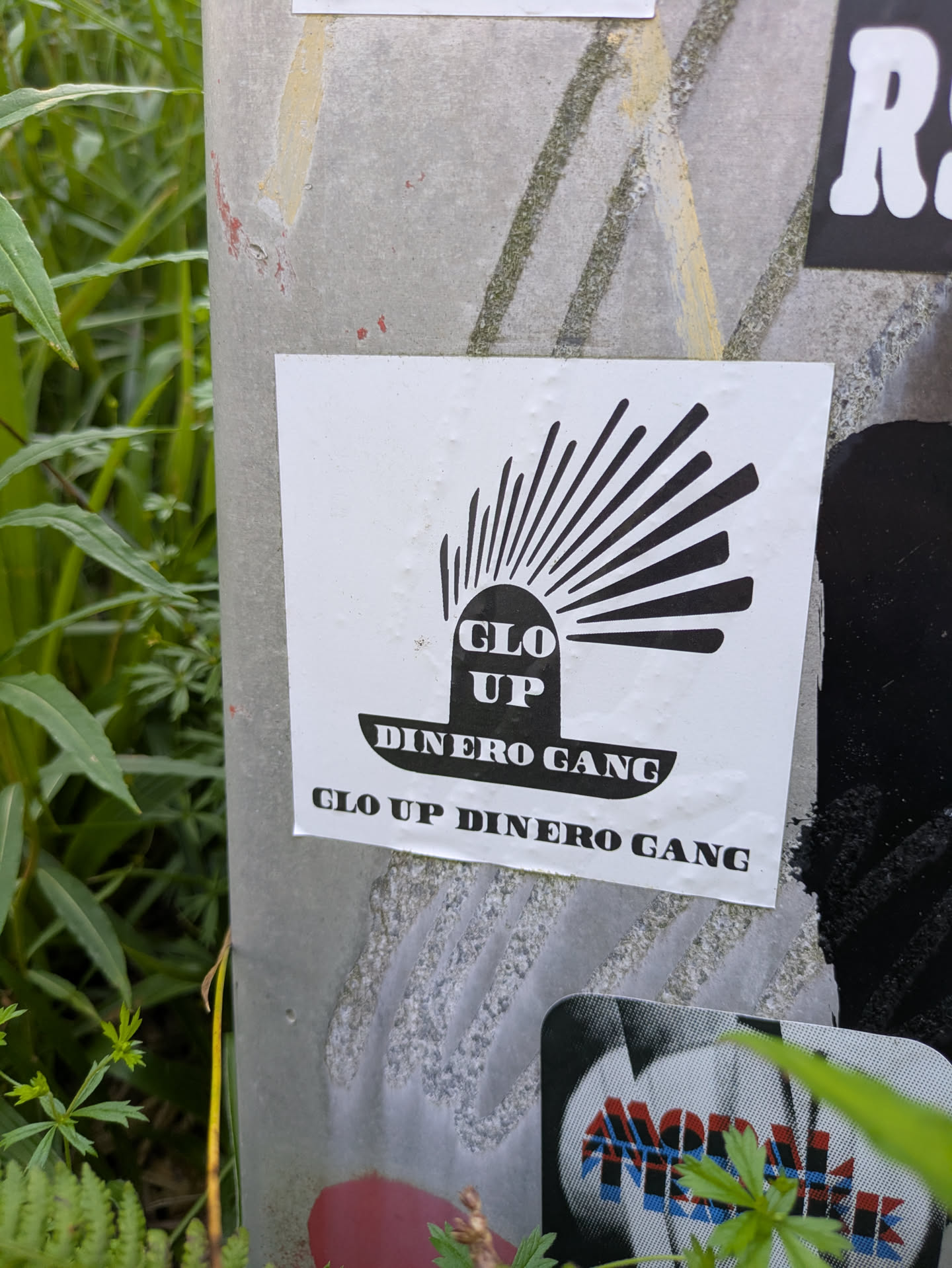 The sticker is white with a central black graphic resembling a sun or a rising symbol with rays. Inside the base of this symbol, the words "GLO UP" are printed vertically. Below the symbol, the words "DINERO GANG" are written horizontally. The full phrase "GLO UP DINERO GANG" is repeated again in black text at the bottom edge of the sticker. The sticker is placed on a light grey pole, with green foliage visible around it.