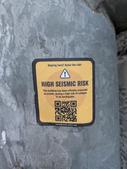 A square sticker with rounded corners on a pole. The sticker is yellow with a black border. The text on the sticker indicates that the building has been officially evaluated as unsafe, posing a high risk of collapse in an earthquake. There is a QR code at the bottom of the sticker and the text "Staying here? Know the risk!"