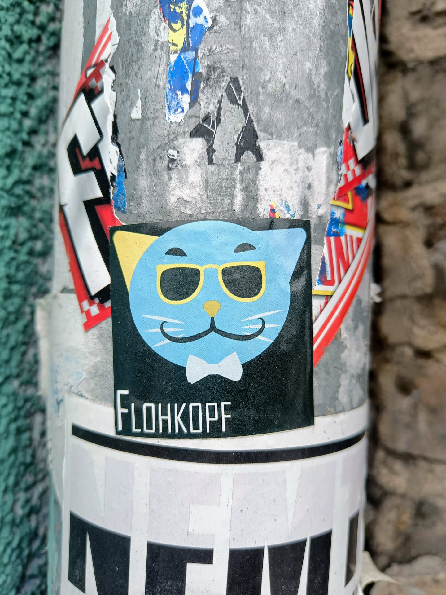 A black sticker with a cartoon illustration of a light blue cat wearing sunglasses and a bow tie. The text 'FLOHKOPF' is visible at the bottom of the sticker. The sticker is affixed to a pole covered in other stickers and graffiti.