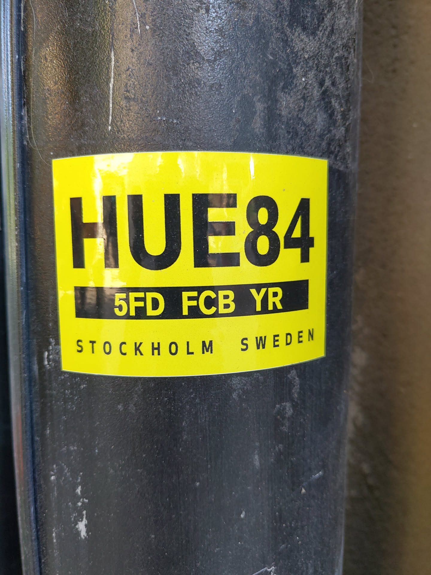 A yellow and black sticker with the text HUE84, 5FD FCB YR, and STOCKHOLM SWEDEN. The sticker is attached to a dark gray cylindrical object.