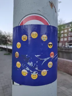 A sticker with various emojis arranged in a circle around a stylized image resembling the European Union flag. The emojis express a range of emotions, from happy to angry. The sticker is affixed to a grey pole.