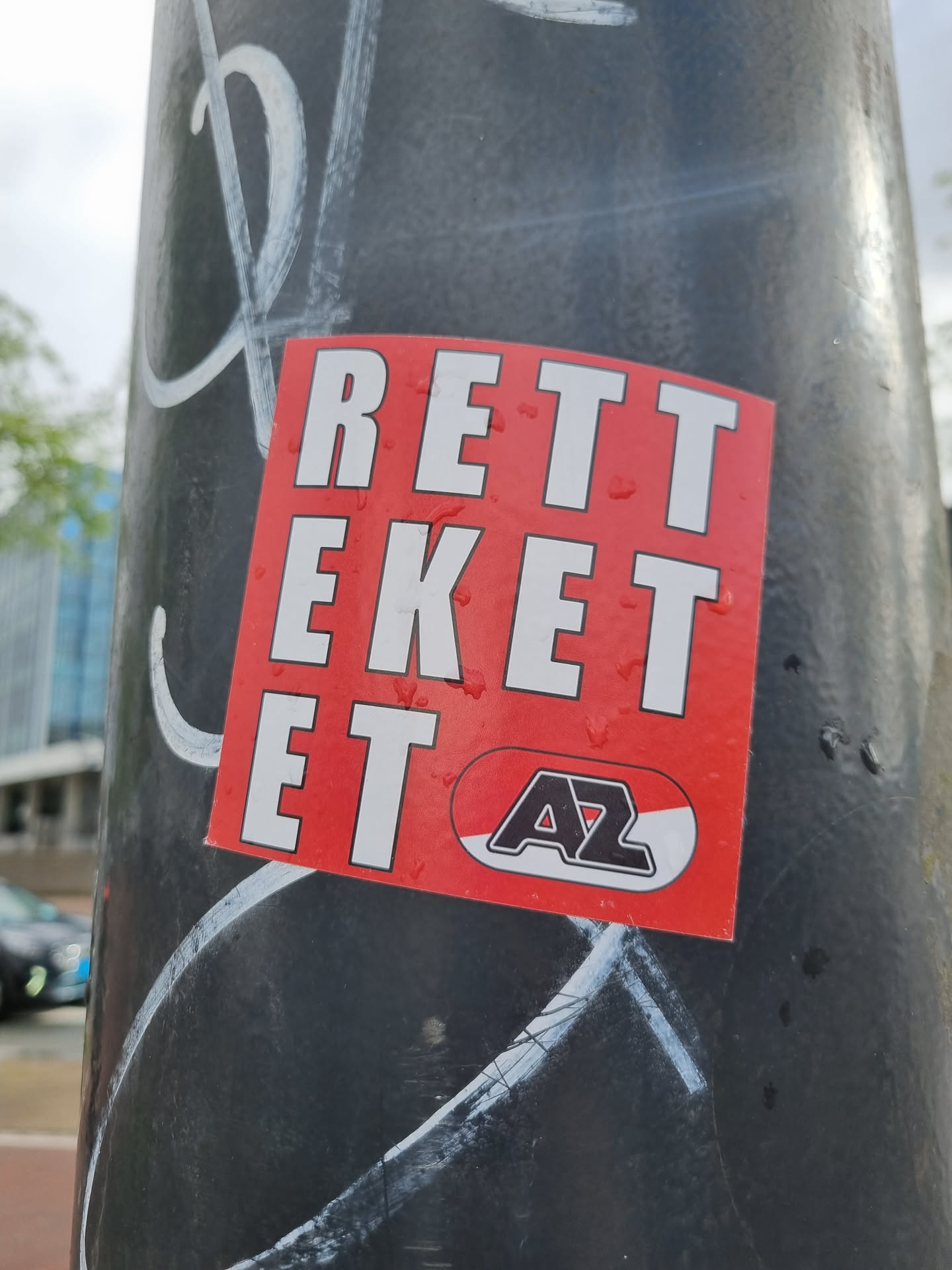 A red sticker with the text "RETTEKEKET" in large white capital letters. There is a smaller logo underneath, possibly related to a sports team, featuring the letters "AZ" in black and white on a red oval background. The sticker is affixed to a dark-colored pole with other graffiti present.  The text appears to be Dutch.