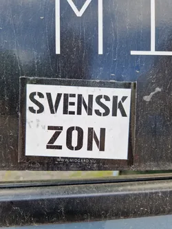 A sticker with the text "SVENSK ZON" (Swedish Zone) and a website address at the bottom. The sticker is white with black stencil-like text. It is affixed to a dark-colored surface.