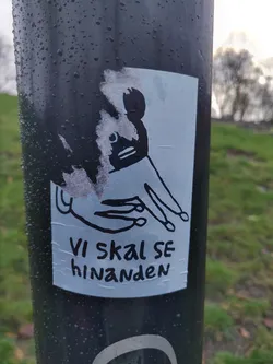 A sticker featuring a simple line drawing of a creature with a hand reaching out. The text below the drawing says "Vi skal se hinanden", which is Danish for "We shall see each other". The sticker is affixed to a dark pole outdoors.
